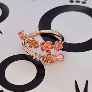 Op de afbeelding: Een roségouden ring met een bloemenontwerp. De ring heeft roze en rode emaille bloemen met kleine edelsteenaccenten. De ring wikkelt zich om de vinger en creëert een unieke en elegante uitstraling. De ring staat tegen een zwart-witte achtergrond.