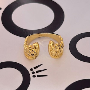 May include: A gold-colored open ring with a detailed floral design on each end. The ring is set against a white background with black circular and linear graphic elements. The ring's interior is stamped with the letters "AH&WA".
