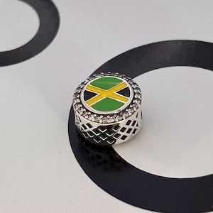 May include: A round charm with the Jamaican flag design. The charm features a green, black, and yellow flag design, surrounded by a ring of clear crystals. The sides of the charm have a lattice pattern.