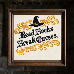 May include: A framed cross-stitch artwork with the words "Read Books Break Curses." It features a black witch's hat, gold embellishments, and black lettering on a cream background, set in a brown wooden frame.