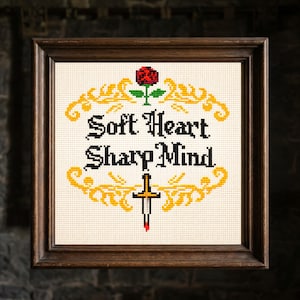 Può includere: Opera d'arte a punto croce in una cornice di legno. Il design presenta le parole "Soft Heart Sharp Mind" in lettere gotiche nere, una rosa rossa e un bordo dorato. Una spada con una goccia rossa è al centro.