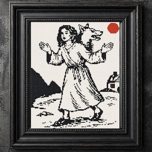 May include: Black framed cross-stitch art featuring a woman with a wolf head, a red sun, and a small house. The artwork is primarily black and white, with the woman in a flowing dress and the wolf head facing right. The background includes mountains and a path.