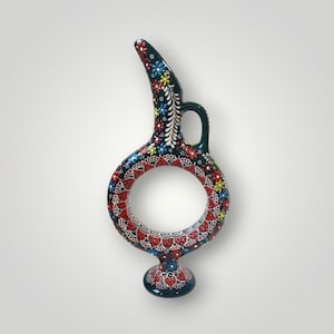 Può includere: Un vaso decorativo in ceramica verde scuro con un design circolare unico. Il vaso è adornato con motivi floreali colorati in rosso, blu, giallo e bianco. Presenta un manico e un beccuccio svasato, mostrando un mix di arte e funzionalità.
