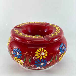 May include: A round, red ashtray with a glossy finish. The ashtray is decorated with hand-painted blue and yellow floral designs. A yellow wavy pattern encircles the rim.