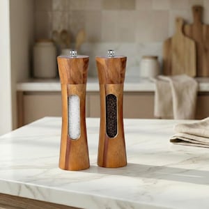 Puede incluir: Dos molinillos de sal y pimienta de madera con acabado de madera natural. Cada molinillo tiene una ventana transparente que muestra el contenido: sal y granos de pimienta. Los molinillos están sobre una encimera blanca.