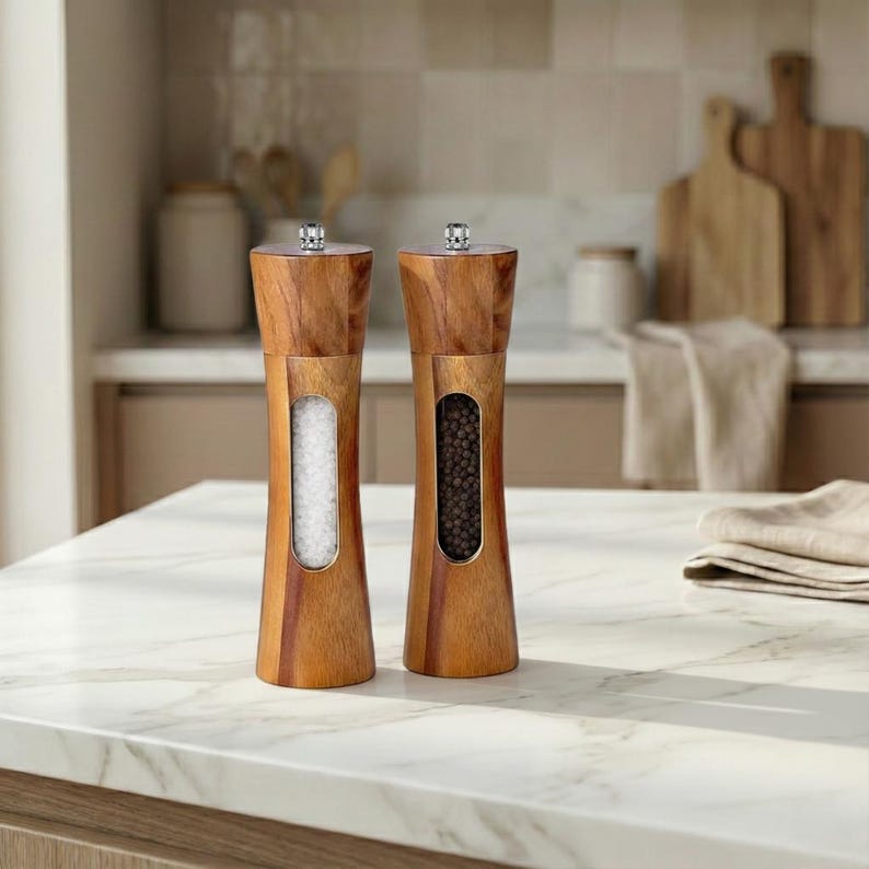 May include: Two wooden salt and pepper grinders with a natural wood finish. Each grinder has a clear window showing the contents: salt and peppercorns. The grinders are on a white countertop.