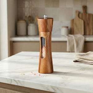 May include: A wooden salt grinder with a clear window displaying pink salt. The grinder has a silver top and a curved design. It is sitting on a white marble countertop, with a neutral background.