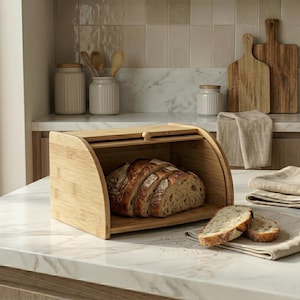 Puede incluir: Una panera de bambú con tapa enrollable, que contiene una hogaza de pan en rodajas. Dos rebanadas de pan descansan sobre un paño de lino. En el fondo se ven accesorios de cocina, como tablas de cortar y tarros.