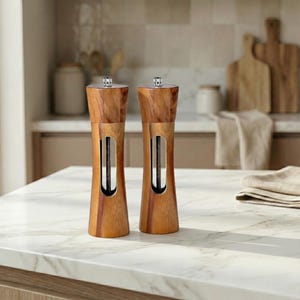 May include: Two wooden salt and pepper grinders with a natural wood finish. Each grinder has a stainless steel accent and a silver top. They are displayed on a white countertop in a kitchen setting.