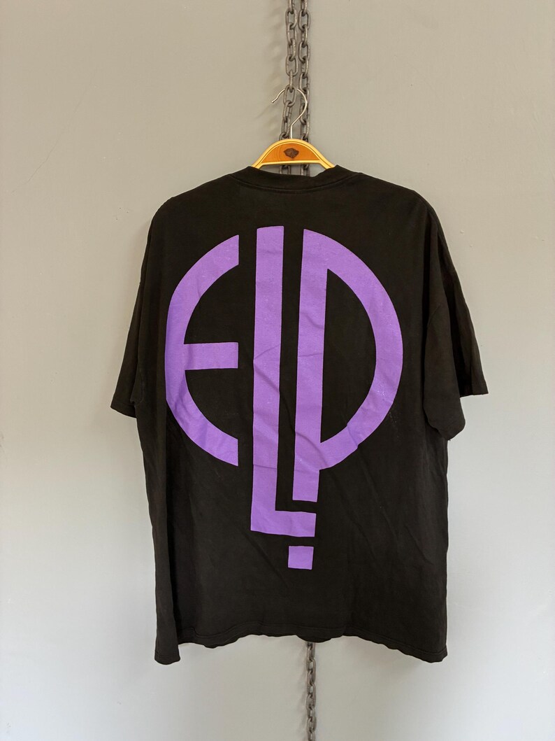 May include: Black t-shirt with a large purple graphic on the back. The graphic is a stylized design of the letters "E", "L", and "P". The shirt is hanging on a wooden hanger.