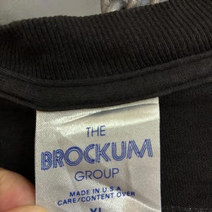 May include: Close-up of a black t-shirt label. The label is white with blue text that reads "THE BROCKUM GROUP" and "MADE IN U.S.A. CARE/CONTENT OVER". The size "XL" is printed at the bottom of the label.