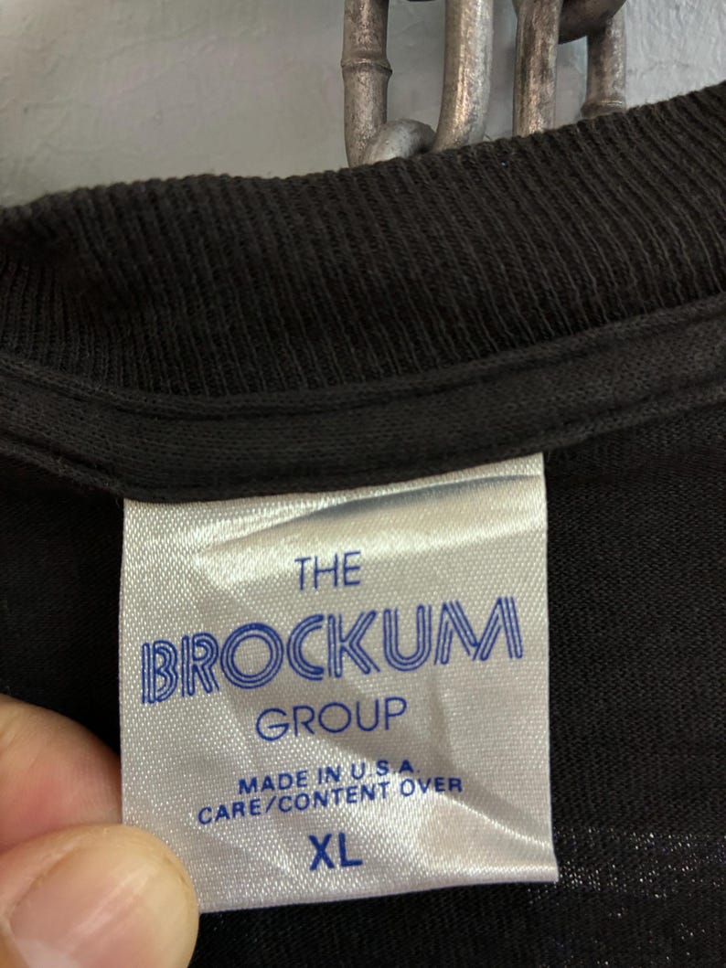 May include: Close-up of a black t-shirt label. The label is white with blue text that reads "THE BROCKUM GROUP" and "MADE IN U.S.A. CARE/CONTENT OVER". The size "XL" is printed at the bottom of the label.
