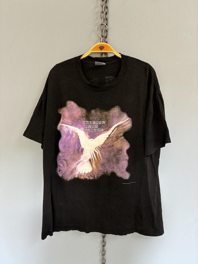 May include: Black t-shirt with a white bird graphic and the text "Jefferson Airplane". The bird has outstretched wings against a purple and brown background. The t-shirt is hanging on a gold hanger.