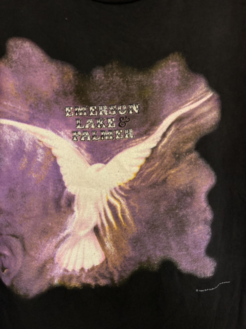 May include: Black t-shirt with a white dove graphic, wings spread, set against a purple and gold backdrop. The text "EMERSON LAKE & PALMER" is above the dove. A vintage design.