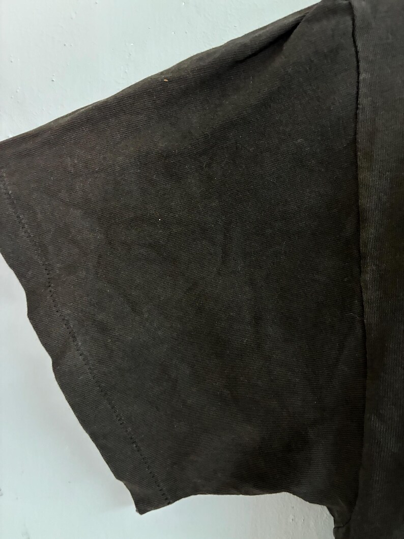 May include: Close-up of a black t-shirt sleeve. The fabric appears to be a soft cotton blend, with visible texture and subtle variations in the dark colour. The sleeve's edge is neatly hemmed with a visible seam.