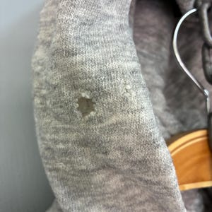 Puede incluir: Primer plano de una sudadera con capucha gris con un agujero en la tela. La sudadera con capucha est&aacute; hecha de un material de punto suave. La capucha est&aacute; colgada en una percha.