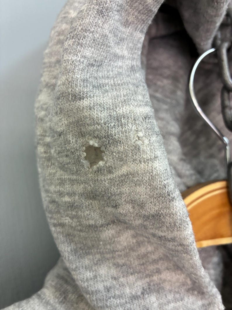 Puede incluir: Primer plano de una sudadera con capucha gris con un agujero en la tela. La sudadera con capucha est&aacute; hecha de un material de punto suave. La capucha est&aacute; colgada en una percha.
