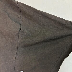 May include: Close-up of a dark brown t-shirt sleeve. The sleeve is made of a textured fabric and shows a seam along the top. The fabric appears slightly faded, with a visible tear near the seam.