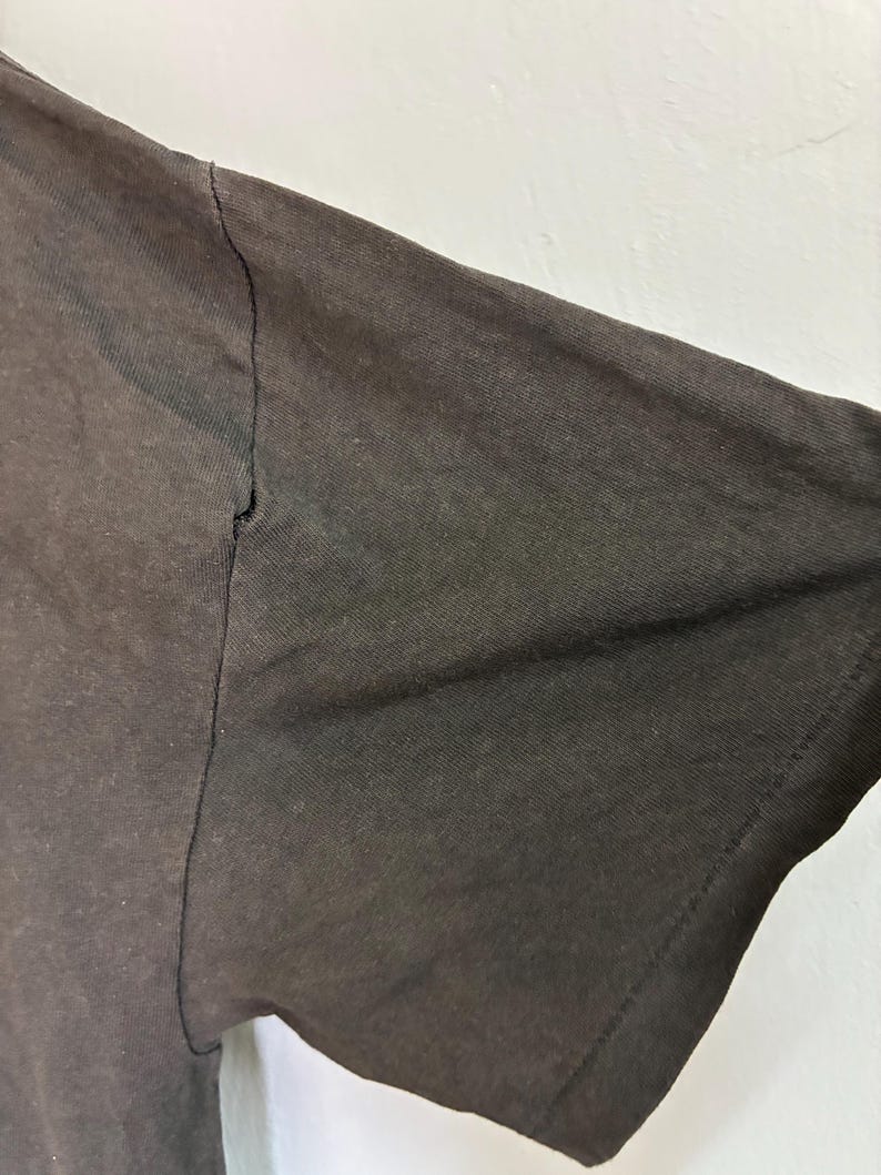 May include: Close-up of a dark brown t-shirt sleeve. The sleeve is made of a textured fabric and shows a seam along the top. The fabric appears slightly faded, with a visible tear near the seam.