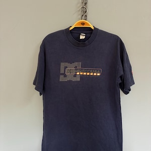 May include: Navy blue t-shirt with a crew neck and short sleeves. The shirt features a logo design in orange and gray, with the text "DCSHOECOUSA" and "ULTIMATE TERRAIN". The shirt is hanging on a wooden hanger.