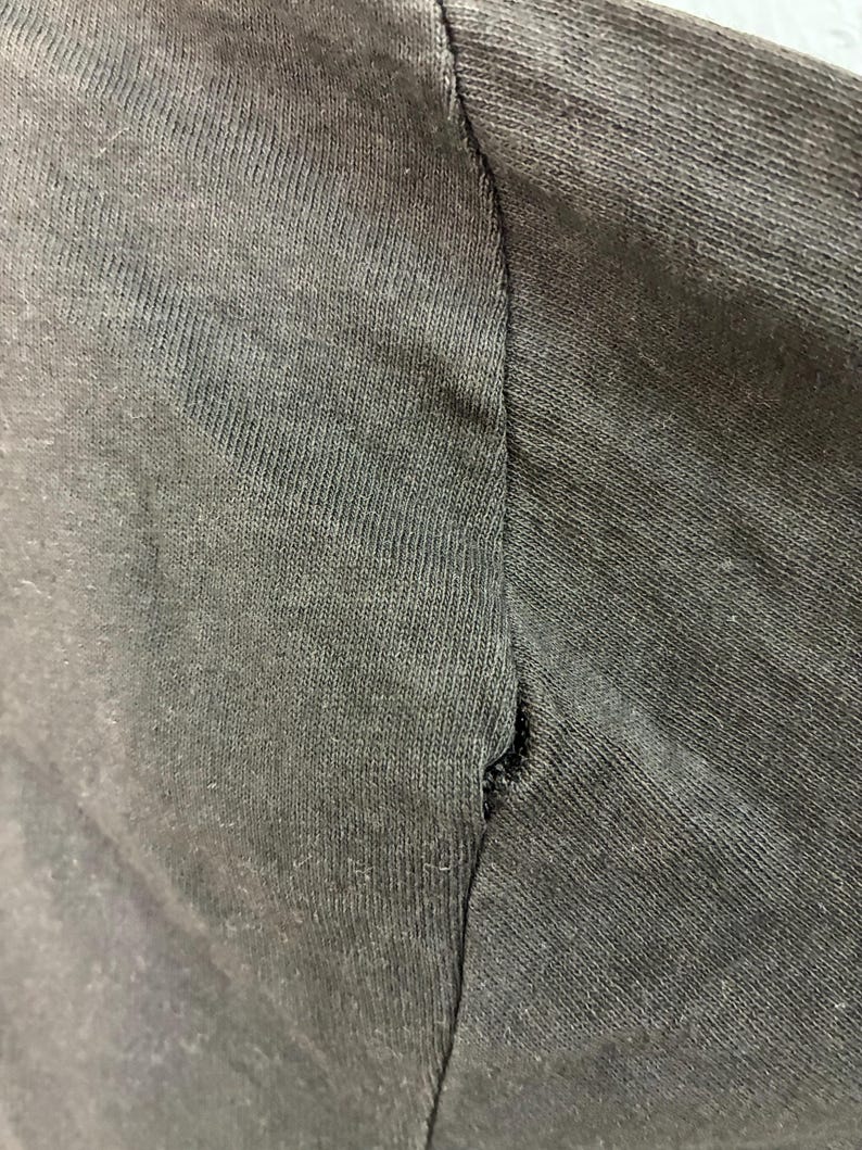 May include: Close-up of a dark grey t-shirt sleeve, showing the seam and fabric texture. The fabric has a ribbed texture, and the seam is slightly frayed.