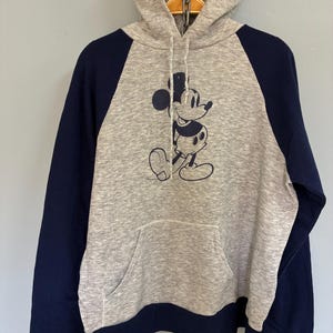 Puede incluir: Sudadera con capucha gris con mangas azul marino y un bolsillo delantero. La sudadera presenta un gr&aacute;fico azul marino de Mickey Mouse. La capucha tiene un cierre de cord&oacute;n. Una parte superior informal y c&oacute;moda.