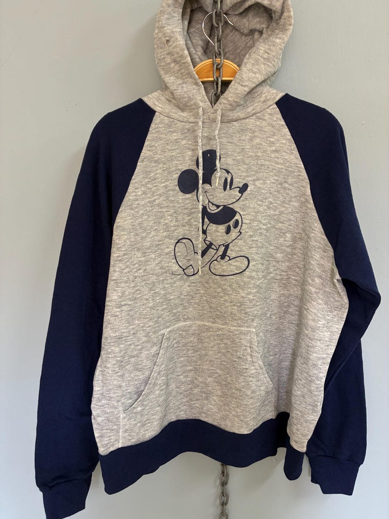 Puede incluir: Sudadera con capucha gris con mangas azul marino y un bolsillo delantero. La sudadera presenta un gr&aacute;fico azul marino de Mickey Mouse. La capucha tiene un cierre de cord&oacute;n. Una parte superior informal y c&oacute;moda.
