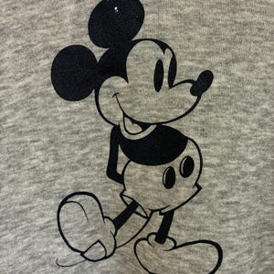 Puede incluir: Una tela gris con un gr&aacute;fico negro de Mickey Mouse. El personaje de dibujos animados se representa en una pose cl&aacute;sica, con sus ic&oacute;nicas orejas grandes, su rostro sonriente y sus guantes blancos. La tela tiene una apariencia texturizada.