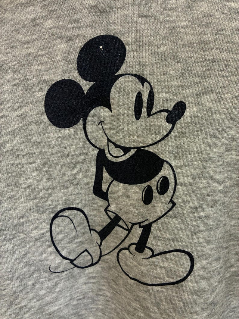 Puede incluir: Una tela gris con un gr&aacute;fico negro de Mickey Mouse. El personaje de dibujos animados se representa en una pose cl&aacute;sica, con sus ic&oacute;nicas orejas grandes, su rostro sonriente y sus guantes blancos. La tela tiene una apariencia texturizada.