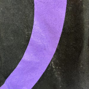 May include: Close-up of black fabric featuring a broad, curved, purple stripe. The stripe has a textured finish and is angled across the image. The fabric shows signs of wear and tear.