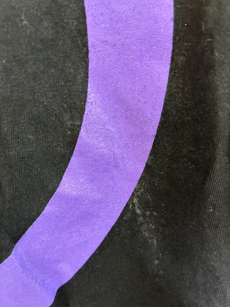 May include: Close-up of black fabric featuring a broad, curved, purple stripe. The stripe has a textured finish and is angled across the image. The fabric shows signs of wear and tear.