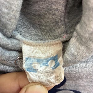 Puede incluir: Primer plano de una sudadera con capucha gris con una etiqueta blanca. La etiqueta tiene las palabras "CASUALS" y "X-LARGE" impresas. La sudadera tiene un gr&aacute;fico de Mickey Mouse.