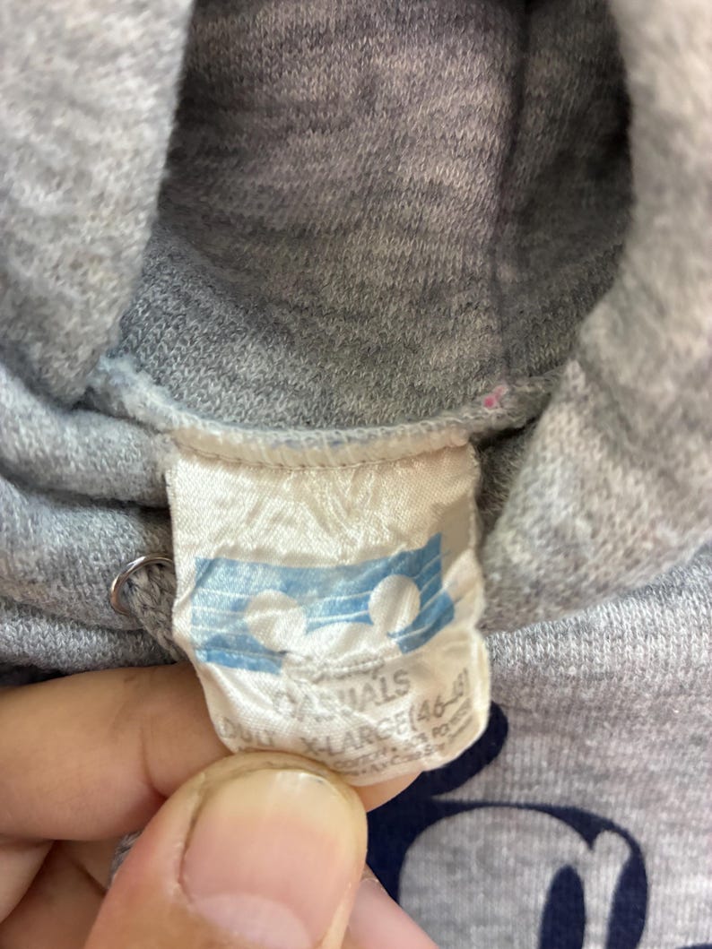 Puede incluir: Primer plano de una sudadera con capucha gris con una etiqueta blanca. La etiqueta tiene las palabras "CASUALS" y "X-LARGE" impresas. La sudadera tiene un gr&aacute;fico de Mickey Mouse.