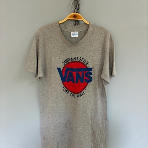以下が含まれることがあります： クルーネックと半袖のグレーのTシャツ。赤と青のVansのロゴが特徴で、上部に「SUNVANS STYLE」、下部に「OFF THE WALL」と書かれています。Tシャツは木製のハンガーに吊るされています。
