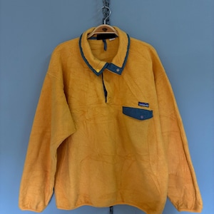May include: A bright yellow long-sleeve fleece pullover featuring a blue collar and a matching blue chest pocket with a snap button closure. The garment includes a partial snap-button placket at the neckline and a small Patagonia brand label.