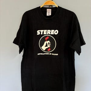 May include: Black t-shirt featuring the word "STEREO" in white, above a circular graphic of a hand holding a red musical note. The text "REVOLUTION IN SOUND" is below the graphic. The shirt is displayed on a wooden hanger.
