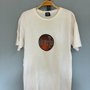May include: White t-shirt with a round graphic featuring the text "silver lab" in a stylized font over an orange and black background. The shirt is hanging on a wooden hanger.