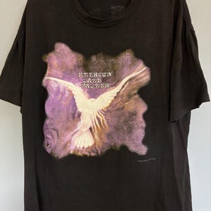 May include: Black t-shirt with a graphic of a white bird with outstretched wings set against a purple and yellow backdrop. The text "EMERSON LAKE & PALMER" is above the bird. A vintage music-themed tee.