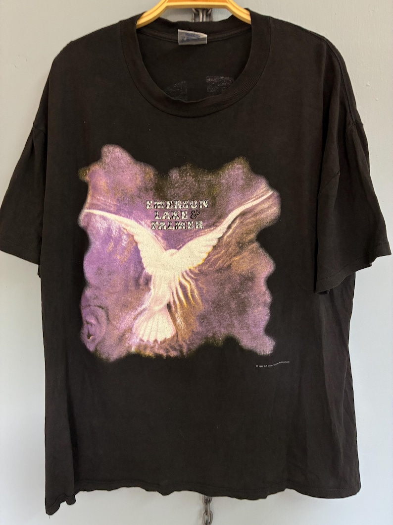 May include: Black t-shirt with a graphic of a white bird with outstretched wings set against a purple and yellow backdrop. The text "EMERSON LAKE & PALMER" is above the bird. A vintage music-themed tee.
