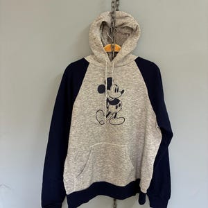 Puede incluir: Una sudadera con capucha gris con mangas azul marino y un bolsillo delantero. La sudadera presenta una ilustraci&oacute;n en blanco y negro de Mickey Mouse. La sudadera con capucha cuelga de una cadena.