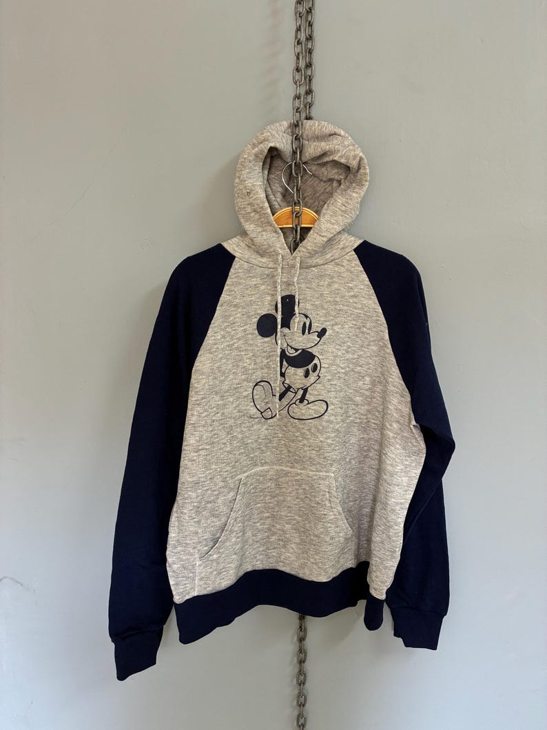 Puede incluir: Una sudadera con capucha gris con mangas azul marino y un bolsillo delantero. La sudadera presenta una ilustraci&oacute;n en blanco y negro de Mickey Mouse. La sudadera con capucha cuelga de una cadena.