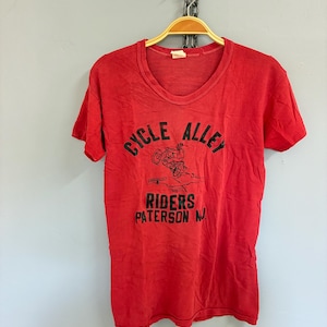 Op de afbeelding: Een rood t-shirt met een ronde hals en korte mouwen. Het shirt toont de tekst "CYCLE ALLEY RIDERS PATERSON N.J." in zwart, samen met een motorfietsafbeelding. Het t-shirt hangt aan een houten hanger.