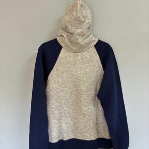 Puede incluir: Una sudadera con capucha gris con mangas y ribetes azul marino. La sudadera con capucha cuelga de una cadena de metal contra una pared gris. La sudadera tiene un estilo cl&aacute;sico y deportivo.