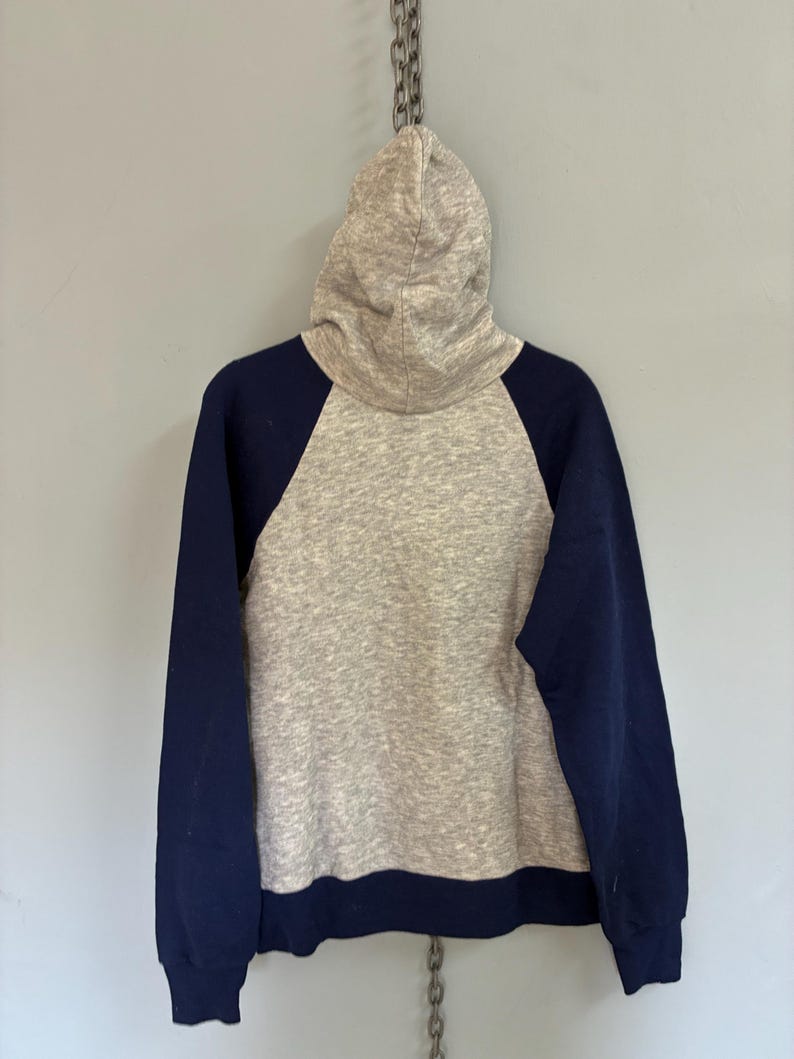 Puede incluir: Una sudadera con capucha gris con mangas y ribetes azul marino. La sudadera con capucha cuelga de una cadena de metal contra una pared gris. La sudadera tiene un estilo cl&aacute;sico y deportivo.