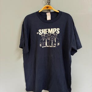 Könnte beinhalten: Marineblaues T-Shirt mit weißer Grafik einer Band und dem Text "The Shemps" über den Bandmitgliedern. Darunter steht der Text "What Dave Wants Means To Me...". Das T-Shirt hängt an einem gelben Kleiderbügel.