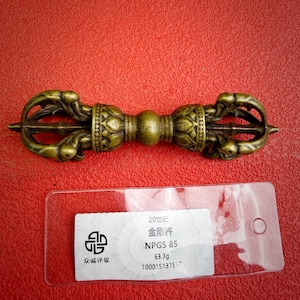 May include: A bronze-colored metal vajra with intricate symmetrical carvings on both ends and a central spherical grip. Below the object, a clear plastic tag displays the text 20世纪, 金刚杵, NPGS 85, 63.3g, and 100015131557, indicating a weight of 63.3 grams.