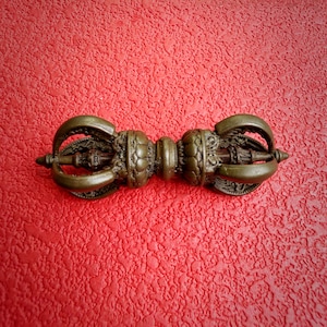 May include: A bronze-colored metal vajra ritual object featuring a central handle with intricate floral-like carvings and symmetrical prongs extending from both ends, creating a balanced, multi-faceted design with a dark, weathered patina finish.