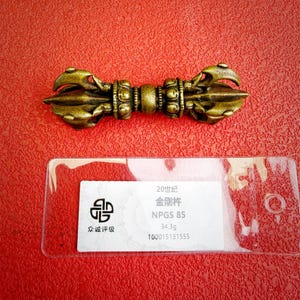 May include: A bronze-coloured metal vajra with intricate symmetrical prongs on both ends and a central grip. Below the object is a clear plastic tag containing the text 20世纪, 金刚杵, NPGS 85, 34.3g, and 100015131555, along with a circular logo.