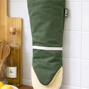 Op de afbeelding: Een groene ovenwant met een witte band en een crèmekleurig, getextureerd interieur. De want hangt aan een houten peg. Het label aan de zijkant luidt "ORLA".