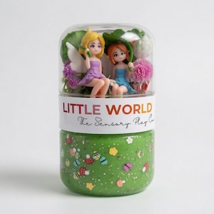 May include: A clear plastic jar containing two small fairy figurines, one in a purple dress and one in a blue dress, surrounded by artificial flowers. A white label reads LITTLE WORLD The Sensory Play Co. The base is filled with green glittery dough.